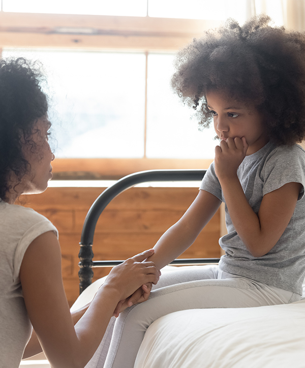 an adult kneels by a child sitting on a bed and holds her hand